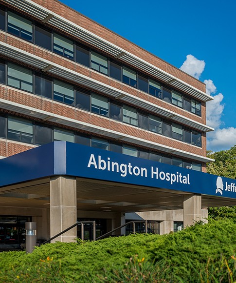 The exterior of a multi-story, brick building with the words “Abington Hospital - Jefferson Health” on a blue backdrop above the entranceway with green shrubbery in front of it.