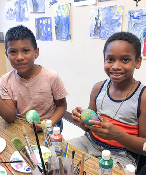 Two children sit at a table covered in paint, paintbrushes and various other art supplies while doing arts and crafts.