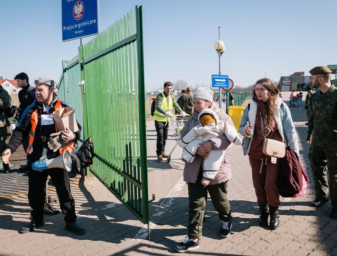 Refugees bundled up in winter coats cross the border through a tall green fence surrounded by border employees in bright vests and camouflage along with a soldier in camouflage.