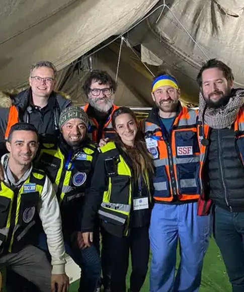Dr. Alex Hajduczok and his team stand inside a large military tent while wearing reflective jackets and other medical gear while standing together and smiling at the camera.