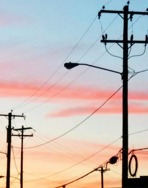 Silhouettes of power lines and streetlights stand against a light blue sky streaked with light pinks, purples, and yellows.