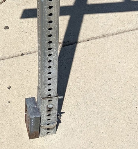 A street signpost on a concrete sidewalk has a rusty box sensor zip-tied around its base as the sun shines and casts a shadow of the signpost on the ground.