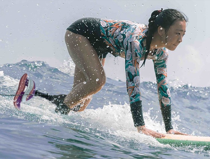 A surfer kneels on their board while catching a wave and wearing a long-sleeved wetsuit top with swim trunks, and a rendering shows the Swell Surf Foot prosthetic on their left leg.