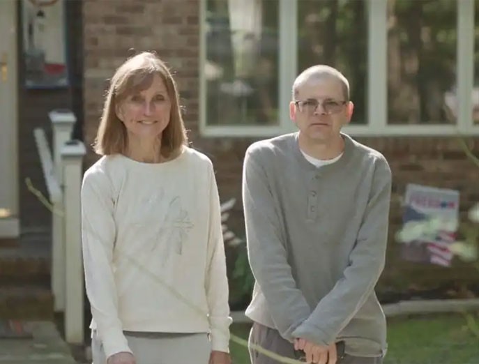 Aaron wears a long-sleeved gray shirt and glasses, leans on a cane, and looks at the camera while standing next to his mother, Holly, who’s wearing a long-sleeved cream top while smiling at the camera.