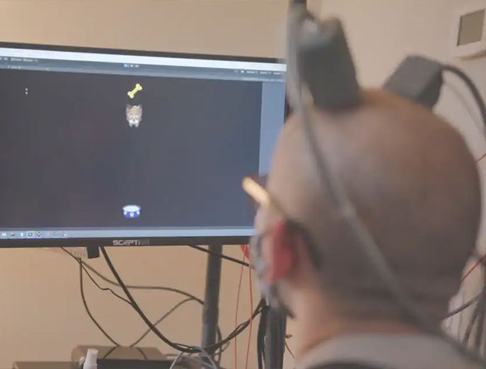 Aaron wears electrodes on his head while looking at a monitor displaying a video game featuring a brown dog, a yellow bone and a blue dog food bowl.