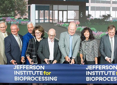 A large group of people in formal attire stands while holding scissors in preparation to cut a large blue ribbon that reads “Jefferson Institute for Bioprocessing.”
