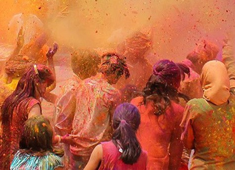 A large group of people dressed in an array of brightly colored clothing have their backs turned to the camera while a colorful powder flies all around in the air.