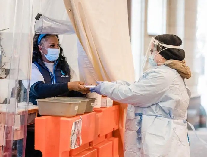 Two medical workers wearing face masks and other forms of personal protective equipment pass an object between them while one stands inside of a tent and the other stands outside of it.