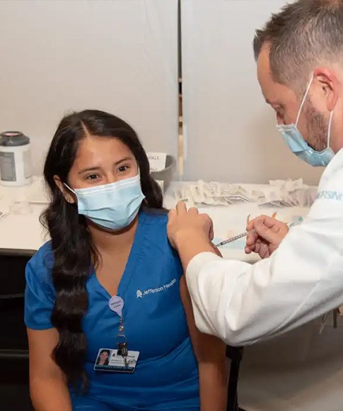 A Jefferson nurse wears blue scrubs, her ID badge and a face mask while receiving the first COVID-19 vaccine at Thomas Jefferson University Hospital from a medical worker wearing a white lab coat and a face mask.