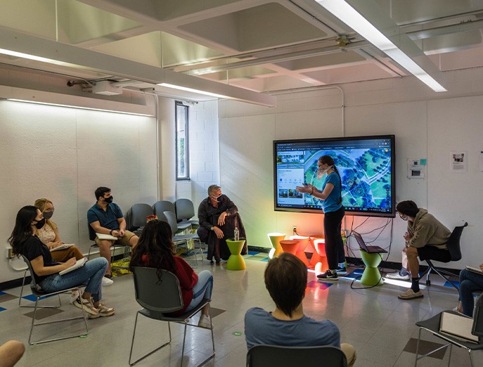 Ivano D’Angella leads an architecture design course in the Architecture and Design Center, where students are spaced out in a circle while wearing face masks and watching one student speak in front of a large screen on the wall. 