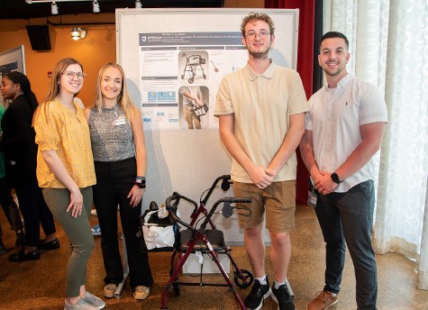 The Roll and Go Basket team stands in front of their project poster and next to the prototype basket on a walker at the 2023 Design Assistive Device Project presentation.